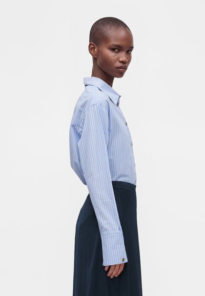 Femme aux cheveux courts portant une chemise à rayures bleu clair boutonnée et une jupe foncée, debout de profil sur un fond blanc uni.