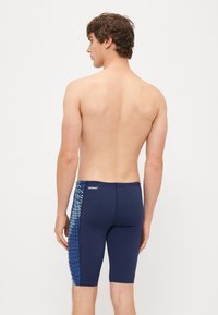 Shorts de bain bleu marine avec une coupe mi-cuisse, présentant un panneau latéral à motifs bleus. Fabriqués en tissu extensible pour le confort et la flexibilité.