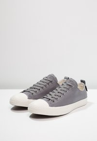 Pár nízkých šedých plátěných tenisek s bílými gumovými podrážkami a špičkami, zašněrované, umístěné na bílé ploše.