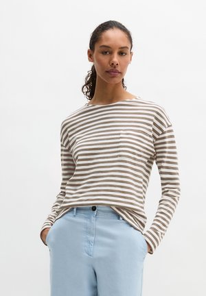 Vrouw met krullend haar in een staart, gekleed in een beige-wit gestreept shirt met lange mouwen en lichtblauwe broek, met handen in de zakken.