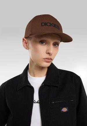 Brun kasket med flad skygge og sort "DICKIES" logo. Kombinationen inkluderer en sort jakke med lynlåslomme og en hvid skjorte under.