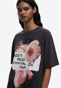 Musta ylisuuria t-paita, jossa on graafinen painatus kasvoista pinkkien kukkien kanssa ja teksti "ÄLÄ USKO KAIKKEA, MITÄ AJATTELET."