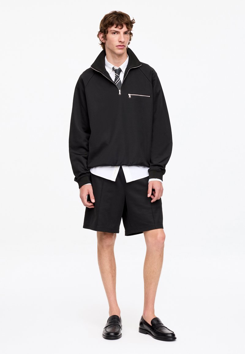 Jeune homme debout, vêtu d'un pull noir zippé, d'une chemise blanche, d'une cravate rayée, d'un short noir et de mocassins noirs sur un fond uni.