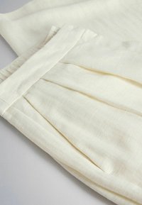 Prim-plan cu un material de culoare crem deschis, cu pliuri moi și cusături vizibile pe o haină, probabil pantaloni sau o fustă.