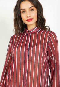 Blusa roja transparente con rayas verticales en blanco y azul marino, con cierre de botones y un cuello clásico. Mangas largas con puños de botón.
