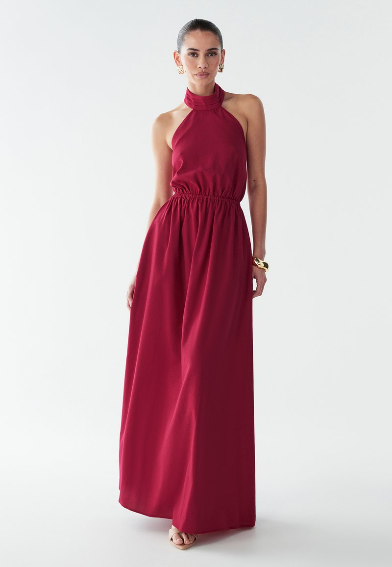 Vestido maxi burdeos con cuello halter, cuerpo ajustado, cintura elástica y falda fluida. Confeccionado con tela suave, presenta una textura lisa y un diseño sencillo.