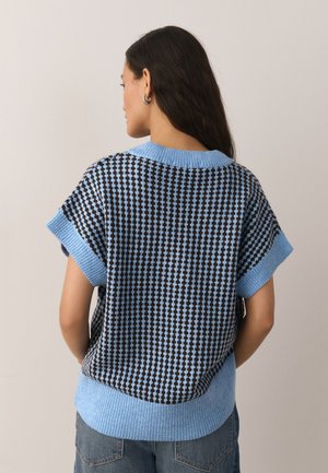 Donna con capelli lunghi e scuri vista di spalle che indossa un gilet a maniche corte blu e nero con motivo e jeans blu.