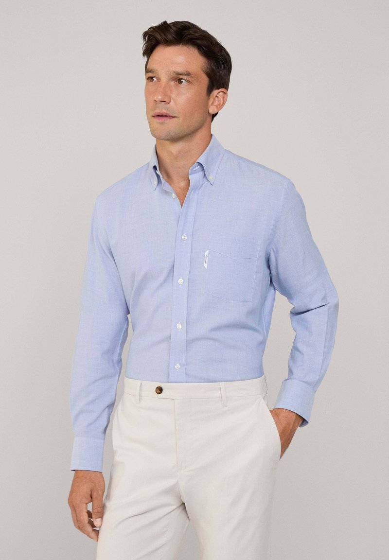 Camisa de botones azul claro con un bolsillo en el pecho, cuello y mangas largas, hecha de tela ligera. Combinada con pantalones color crema.