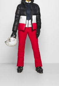 Zwarte pufferjack met rode en witte accenten, gecombineerd met felrode skibroek. Zwarte skibotten en handschoenen, met een witte helm in de hand.