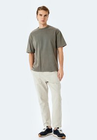 Camiseta oversize verde oliva, pantalones tapered beige pálido y zapatillas negras con cordones blancos. El modelo se presenta en una pose relajada.