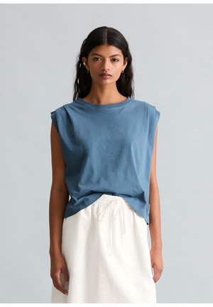 Woman with dark hair wearing a blue sleeveless top and white skirt, standing against a plain light gray background.