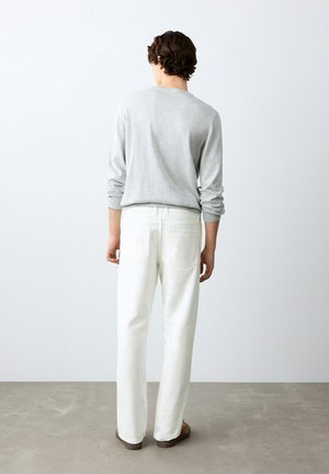 Uomo con capelli ricci che indossa un maglione grigio chiaro e pantaloni bianchi, in piedi di fronte a una parete bianca semplice su un pavimento grigio.