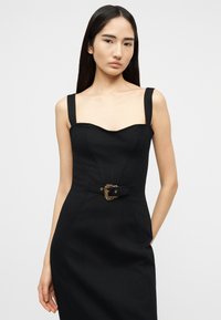 Νεαρή γυναίκα με μακριά σκούρα μαλλιά, ντυμένη με στενό μαύρο φόρεμα με φαρδιές τιράντες και διακοσμητική αγκράφα ζώνης, που στέκεται μπροστά σε απλό φόντο.