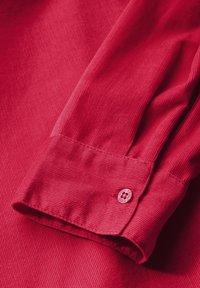 Détail d'une chemise en velours côtelé rouge, avec une texture lisse, un poignet replié et un accent de bouton rouge avec un motif floral.