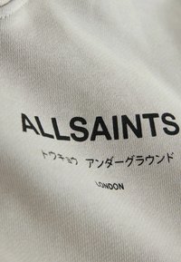 Nahaufnahme eines beigen Stoffes mit "ALLSAINTS" in fetten schwarzen Buchstaben, darunter japanischer Text und darunter in kleinerer Schrift "LONDON".