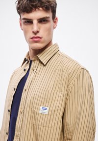 Tan rjavo srajca s črtnim vzorcem, gumbi na sprednji strani, z levim žepom na prsih in označeno etiketo, izdelana iz lahkega materiala, z ovratnikom in črnimi gumbi.