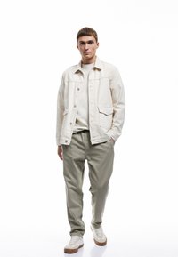 Lys beige denimjakke med to forlommer, parret med olivengrønne bukser og hvide sneakers med brune såler. En ensfarvet hvid t-shirt under.