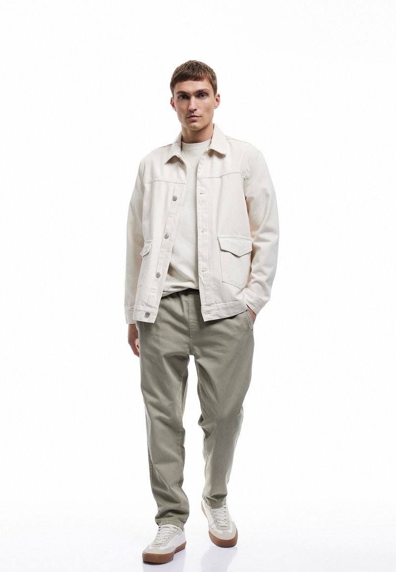 Lys beige denimjakke med to forlommer, parret med olivengrønne bukser og hvide sneakers med brune såler. En ensfarvet hvid t-shirt under.