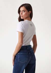Hvid kortærmet t-shirt med round neckline, der har trykt tekst på den øverste ryg. Kombineret med højtaljede mørkeblå jeans.