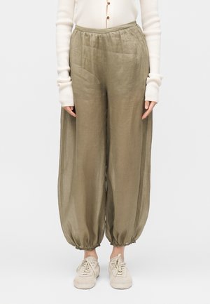 Kobieta nosząca luźne oliwkowo-zielone spodnie ze ściągaczami przy kostkach, biały, długi rękaw z guzikami i beżowe sznurowane buty.