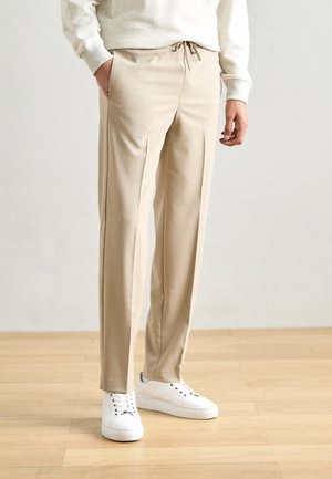 Beige broek met een trekkoord in de taille, zijzakken en een recht gesneden ontwerp. G gedragen met witte sneakers tegen een neutrale achtergrond.