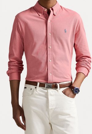 Homme portant une chemise rose à manches longues avec des boutons, un pantalon blanc, une ceinture marron et une montre bleue, la main dans la poche.