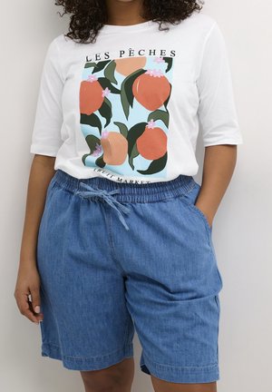 Person wearing white shirt with peach illustrations and text, paired with blue denim drawstring shorts with hands in pockets.