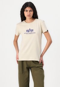 Smėlio spalvos medvilnės trumpomis rankovėmis marškinėliai, kuriuose yra mėlynas grafikų logotipas "ALPHA INDUSTRIES" ant krūtinės. Dėvimi su žaliais cargo kelnėmis.