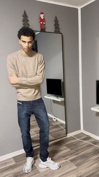 Homme en pull beige et jean, debout, les bras croisés dans une pièce avec un miroir mural, une télévision sur une étagère flottante, et des décorations de Noël.