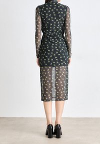 Eine Frau trägt ein durchsichtiges schwarzes Kleid mit langen Ärmeln und grünen Augenmustern, kombiniert mit schwarzen High Heels, und steht vor einer weißen Wand.