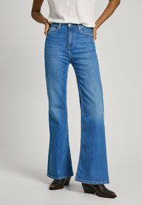 Jeans en denim bleu taille haute évasés avec une texture lisse et un léger délavage, associés à des bottes à talons noires.