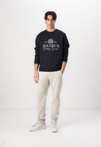 Sudadera navy con texto del logo en blanco, pantalones cargo crema y zapatillas blancas. Textura suave, cuello redondo, mangas largas, corte relajado.