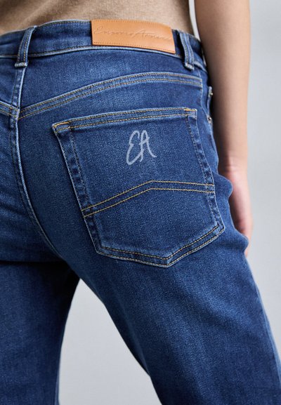 Σκούρες μπλε τζιν παντελόνες με υφή, διαθέτοντας μια πίσω τσέπη με ένα διακριτικό λογότυπο "EA" και αντίθετη κίτρινη ραφή.