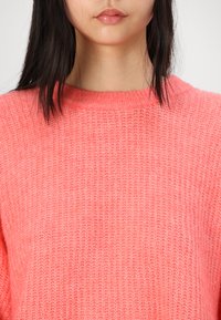 Koral pink strikkegenser med en teksturert, ribbestrikket mønster og rund hals. Stoffet ser mykt ut, med litt løse ermer.