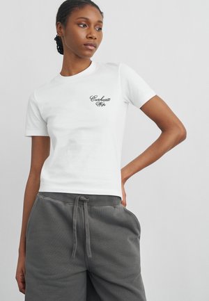 Joven con el pelo trenzado lleva una camiseta blanca de Carhartt Wip y pantalones de chándal grises, posando con una mano en la cadera.