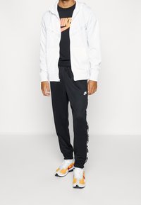 Biała bluza z zamkiem, czarny t-shirt z logo Nike, czarne spodnie dresowe z białymi paskami po bokach oraz wielokolorowe buty do biegania.