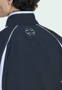 Melna jaka ar augstu apkakli, baltām akcentiem un apaļu logo uz muguras. Gluda auduma tekstūra un sportisks dizains komfortam.