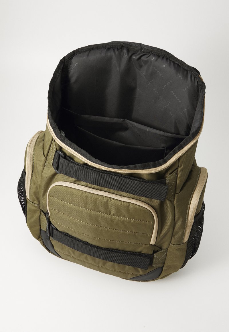 Borsa verde oliva con una forma strutturata, frontale imbottito, spallacci regolabili e interno nero. Presenta diverse tasche e dettagli in rete.