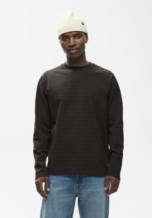 Ung mand iført en mørk stribet langærmet skjorte, lyseblå jeans og en off-white strikket hue på en ensfarvet hvid baggrund.