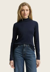 Femme aux longs cheveux blonds, portant un col roulé bleu marine et un jean taille haute bleu, se tenant devant un fond beige uni.