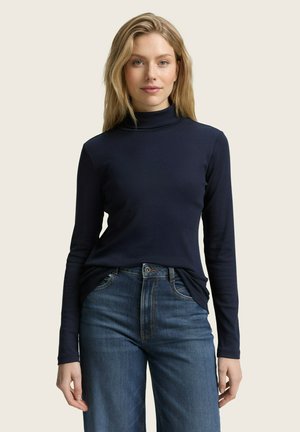 Femme aux longs cheveux blonds, portant un col roulé bleu marine et un jean taille haute bleu, se tenant devant un fond beige uni.
