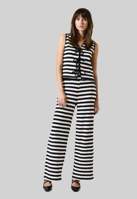 Zwart-wit gestreepte gebreide jumpsuit met een mouwloos ontwerp, voorzien van een knoopsluiting met strikjes en wijde broekspijpen. Aangekleed met zwarte afwerking.