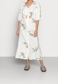 Robe en lin blanc à motifs floraux verts, décolleté en V, manches courtes bouffantes, taille nouée et boutons sur le devant ; associée à des sandales grises.