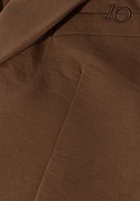 Brązowa tkanina sztruksowa o prążkowanej fakturze, z haftowanym detalem "10". Projekt obejmuje ostre kąty i wyraźne linie.