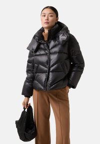 Zwarte pufferjack met een hoge kraag, voorzien van een geborduurd ontwerp, een ritssluiting aan de voorkant en twee zijzakken. Vergezeld van een zwarte tas.