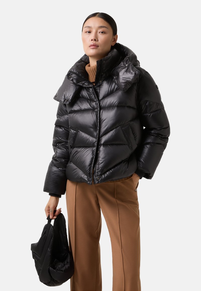 Zwarte pufferjack met een hoge kraag, voorzien van een geborduurd ontwerp, een ritssluiting aan de voorkant en twee zijzakken. Vergezeld van een zwarte tas.