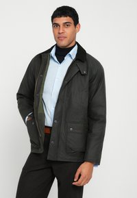 Tmavě zelená bunda z odolné látky s manžetovým límcem, patentními knoflíky a předními kapsami, nošená přes světle modrou košili.