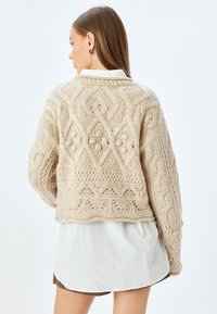 Maglione beige a cima corta con motivo a maglia testurizzato, caratterizzato da design a treccia e a diamante, indossato sopra una camicia bianca. Vestibilità ampia e fondo a coste.