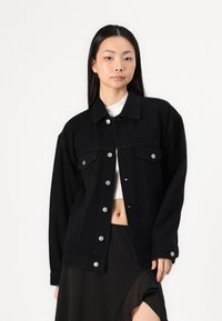Čierna denimová bunda so striebornými gombíkmi, s golierom, dvoma prednými vreckami a voľným strihom. Nosená cez biely crop top.
