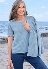 Glunderende vrouw met lang grijs haar die aan de zee staat, gekleed in een lichtblauw structuur T-shirt met korte mouwen, met een bijpassende knooptrui over haar schouder.